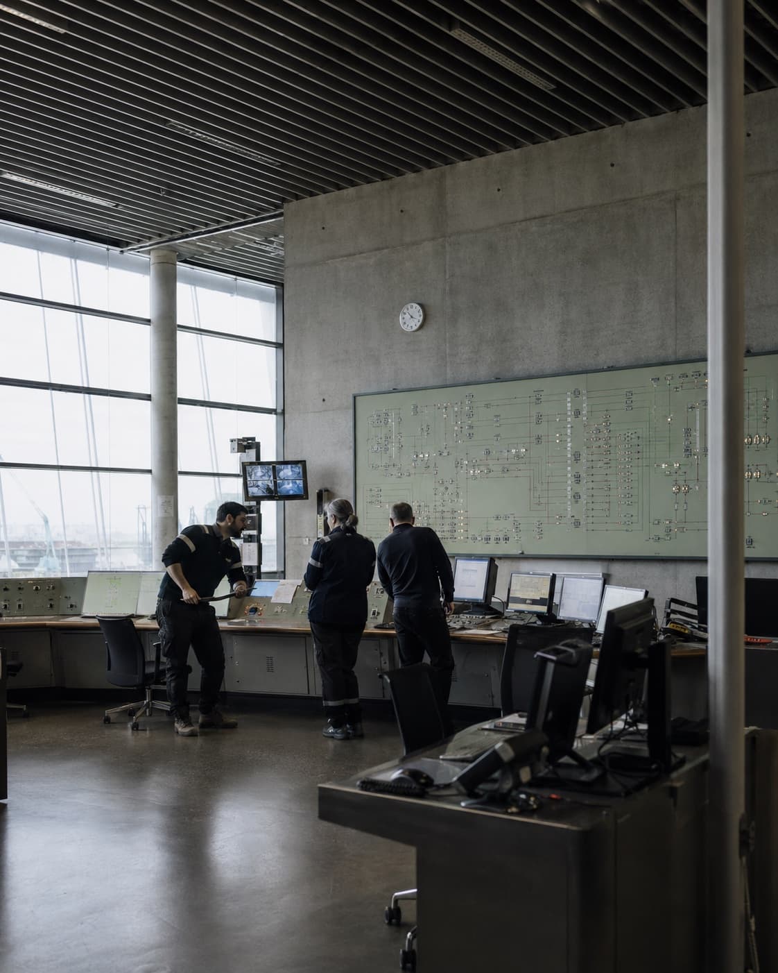 Operators reviewing system state at the consoles of a control room, beneath a wall-sized operational schematic